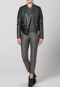 Zelená kožená motocyklová bunda s prošívanými černými ramenními panely, zlatými zipy, spárovaná s vzorovanými šedými kalhotami a černými kotníkovými botami.