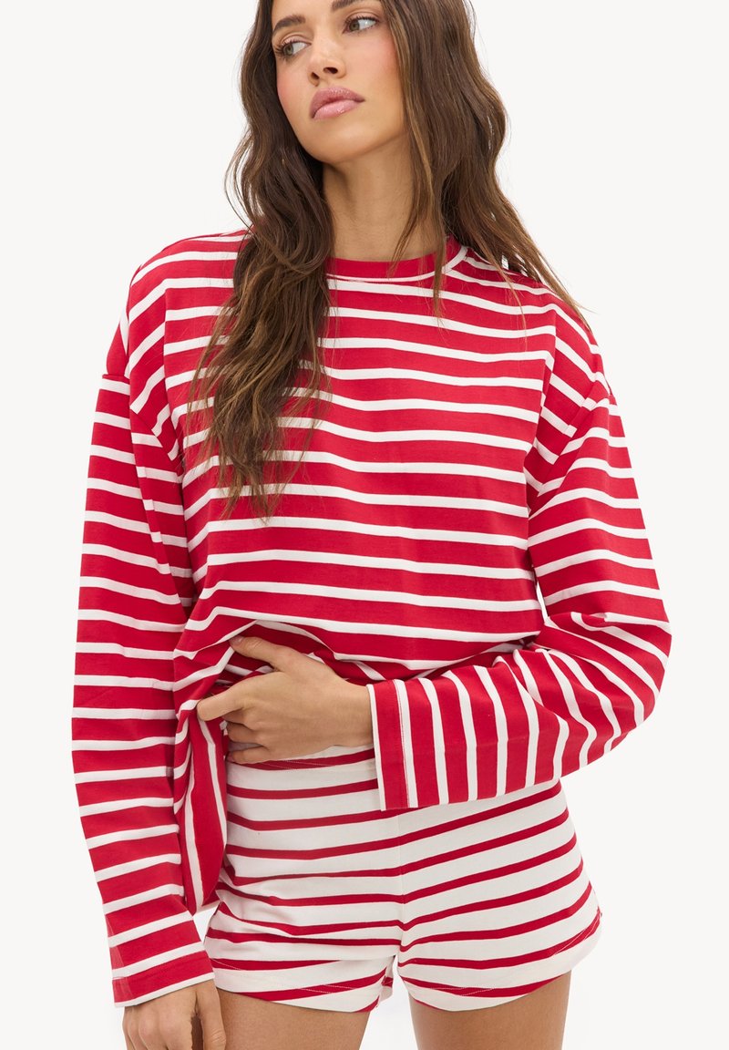 Femme aux longs cheveux bruns portant un haut à manches longues rayé horizontalement rouge et blanc et un short, regardant sur le côté, main sur la taille.