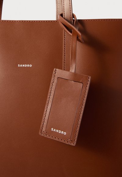 Brun läder tote-väska med en slät yta, med en rektangulär bagagetagg och präglat varumärke "SANDRO" i vitt.