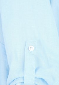 Light blue linen shirt detail featuring a buttoned cuff with a single white button and visible stitching. Textured fabric with a soft appearance.
