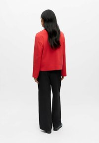 Punane lühike bleiser pikkade varrukatega, millele on lisatud mustad laia lõikega püksid, nähtavad tagant valge tausta ees.