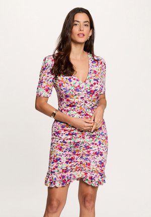 Mujer con un vestido ajustado, fruncido, de flores multicolores, con mangas cortas y bajo con volantes, posando con las manos juntas a la altura de la cintura.