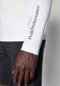 Hvit langermet skjorte med "Peak Performance" trykket i svart på høyre erme, paret med mørke shorts, viser en teksturert hånd.