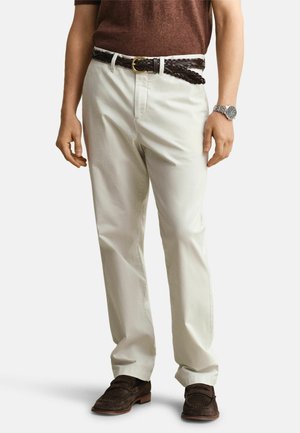 Man wearing cream-colored pants, brown woven belt, brown loafers, brown shirt, and silver wristwatch, standing with relaxed posture.
