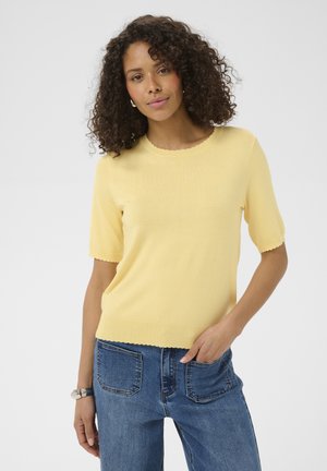 Kobieta o kręconych włosach, ubrana w żółty sweter z krótkim rękawem i niebieskie dżinsy, z ręką spoczywającą w kieszeni, na jednolitym tle.