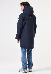 Marineblauer Parka mit Kapuze und langem Schnitt, mit glatter Textur und Logo-Detail am Ärmel. Getragen über hellblauen Jeans.