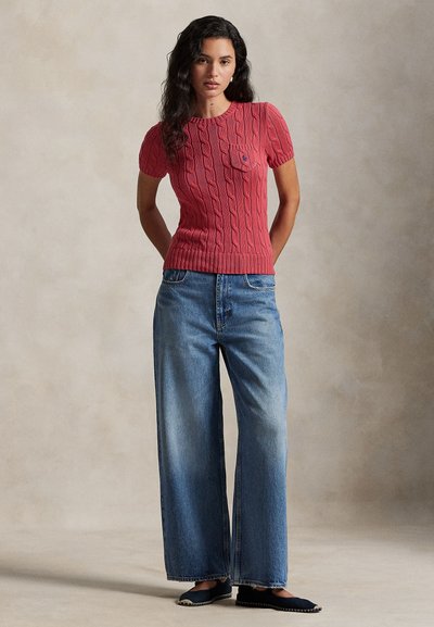 Czerwony, krótki sweter z dzianiny z warkoczowym wzorem i przednią kieszenią, zestawiony z szerokimi, niebieskimi dżinsami i ciemnymi butami.