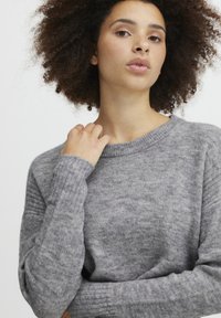 Pull gris en tricot doux avec des poignets et un col côtelés. Présente une coupe décontractée et une surface texturée avec de subtiles variations de teinte.