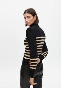 Pulover s ovratnikom u crno-zlatnim prugama, prilagođenog kroja, rebrasti manžeti, mekane teksture, uparen s crnim kožnim hlačama.