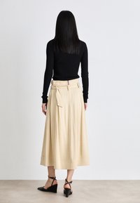 Jupe midi beige avec un détail de taille nouée, fabriquée à partir d'un tissu lisse. Portée avec un haut ajusté à manches longues noir et des chaussures à talons compensés noires.