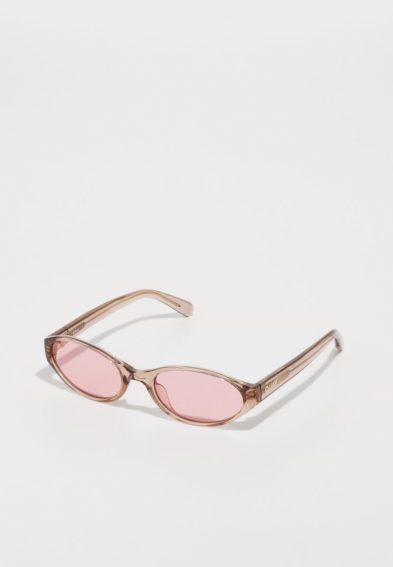 Lunettes de soleil avec une monture marron translucide, des verres ovales teintés de rose, et le nom de la marque "QUAY" embossé sur la branche.