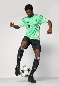 Zelený dres Liverpool FC s čiernymi šortkami, s logom Standard Chartered. Čierne futbalové kopačky s textúrovanou podrážkou a futbalová lopta s hviezdičkovými vzormi.