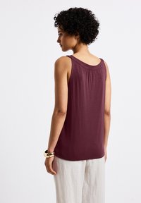 Top sin mangas en color marsala, tejido ligero, escote redondo, pliegues en la parte posterior, combinado con pantalones de lino claros y pulsera de oro.