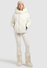 Witte gewatteerde pufferjack met een grote capuchon, in combinatie met witte leggings en kniehoge sneeuwlaarzen met veters. Neutraal gebreide beanie inbegrepen.