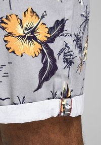 Šedé květinové šortky s žlutými vzory hibiskusu a palmového listu, s bílým lemem a kontrastním stehem na spodním okraji.
