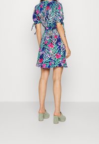 Květinové zavinovací šaty v námořnické modři s růžovými a zelenými vzory, krátké nabírané rukávy a detaily na zavazování. Spárováno se zelenými lodičkami na vysokém podpatku.