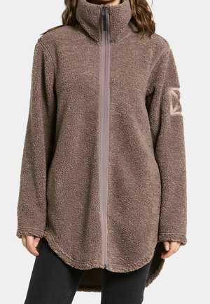 Vrouw draagt een bruin fleecejack met hoge kraag, volledige ritssluiting, lange mouwen en een verlengde, gebogen zoom over een zwarte broek.