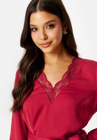 Robe rossa con scollatura a V in pizzo, maniche lunghe e vita legata. La texture del tessuto è liscia e i dettagli in pizzo intricati aggiungono eleganza.