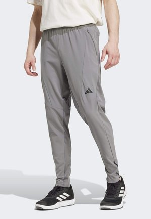 Man die grijze sportbroek met een ritszak draagt en zwarte Adidas-sneakers met witte strepen, staand op een lichte achtergrond.