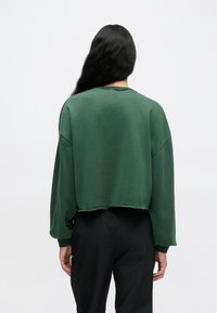 Πράσινο κοντό φούτερ με μακριά μανίκια. Μαλακή υφή, στρογγυλή λαιμόκοψη και ανεπεξέργαστο τελείωμα. Συνδυάζεται με μαύρο παντελόνι, επιδεικνύοντας άνετη εφαρμογή.