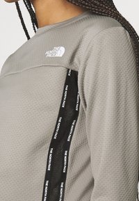Maglia grigia a maniche lunghe con texture, scollo rotondo, caratterizzata da un logo bianco e una striscia laterale nera con testo ripetuto "THE NORTH FACE".
