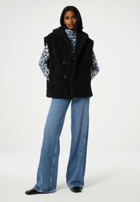 Zwarte gewatteerde bodywarmer met een hoge kraag en dubbele knoopsluiting, gedragen over een bloemenprint lange mouw top, gecombineerd met een wijde blauwe spijkerbroek.