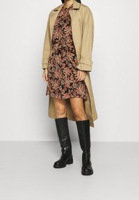 Beige trench coat, kukka-aiheinen ruskea mekko oranssilla ja kermalla painatuksella, sekä mustat polvipituiset saappaat paksulla pohjalla.