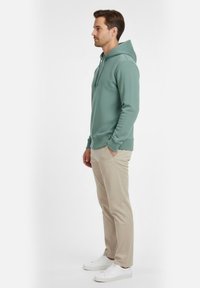 Sudadera con capucha en verde claro, hecha de tela suave, con un bolsillo delantero y puños acanalados; combinada con pantalones beige y zapatillas blancas.
