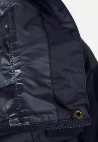 Intérieur de veste bleu marine avec un tissu lisse et brillant, un panneau texturé avec des coutures et un détail de bouton-pression en bronze.