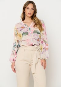 Blusa floreale con maniche lunghe e trasparenti di colore rosa chiaro, caratterizzata da grandi fiori gialli e viola, abbinata a pantaloni beige a vita alta.