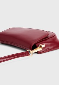 Gladde rode leren polspouch met een goudkleurige sluiting en een afneembare strap tegen een effen lichtgrijze achtergrond.