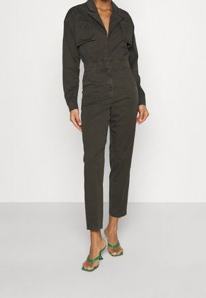 Kobieta ubrana w ciemnooliwkowy kombinezon z długim rękawem i przednimi kieszeniami oraz zielone, odkryte buty na niskim obcasie, stojąca na tle jednolitego białego tła.