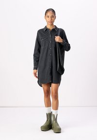 Black denim shirt dress with long sleeves, button front, chest pockets, and curved hem. Paired with green ankle booties and white socks.
