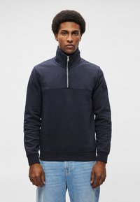 Felpa con zip color navy e colletto alto, realizzata in materiale misto cotone, con polsini e orlo a coste, caratterizzata da una striscia orizzontale sul petto.