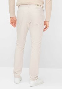 Mann trägt beige Hose und hellen Pullover, steht mit entspannten Händen an den Seiten, weiße Turnschuhe sichtbar vor einfachem Hintergrund.