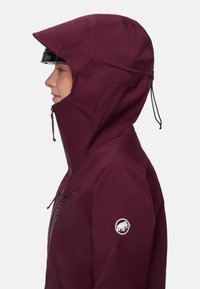 Vinröd vattentät jacka med en åtsittande huva, framdragkedja och justerbar dragsko. Har en logotypmärkning på ärmen. Slät textur.