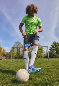 Green PUMA athletic shirt, black shorts with blue accents, white socks, and blue cleats on grass with a weathered soccer ball nearby.