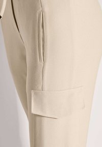 Beige gestructureerde stoffen broek met zijzak en lagere klepzak op de dij.