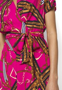Zavinovací šaty v jasně růžové látce se vzorem hnědých řetězů a popruhů, s uvázaným pasem a krátkými rukávy, které zdobí odvážné barvy a tvary.