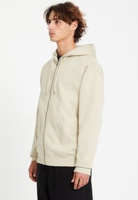 Beige Kapuzenjacke mit Reißverschluss aus weichem Stoff, mit großer Kapuze, Fronttaschen und gerippten Bündchen. Minimalistisches Logo auf der Brust.