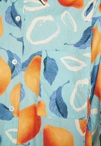 Tissu bleu clair avec motifs de poires orange et feuilles bleu foncé, avec des boutons blancs et une poche poitrine carrée.