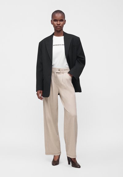 Femme portant un blazer noir oversize, un T-shirt blanc, un pantalon beige à jambes larges et des chaussures marron à talons hauts, debout avec une main dans la poche.