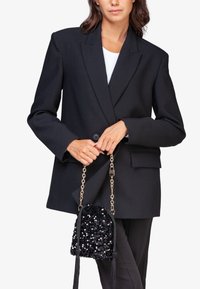 Blazer negro de gran tamaño con un solo botón, combinado con un pequeño bolso negro con lentejuelas que tiene una cadena dorada y borlas.