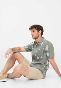 Camicia verde a maniche corte con motivo floreale bianco, abbinata a pantaloni corti beige chiaro e sneakers beige, seduto contro uno sfondo semplice.