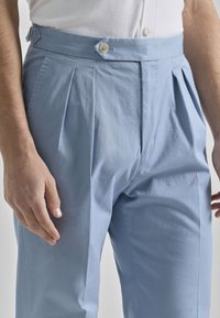Pantalones plegados de color azul claro con una cinturilla que presenta dos botones y una cremallera lateral. La tela es suave con un acabado mate.