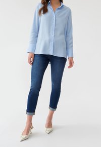 Camisa de botones azul claro de manga larga, combinada con jeans azul oscuro y tacones de punta crema.