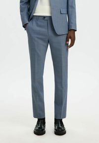 Pantalon bleu clair sur mesure en tissu texturé, avec un devant plat, des poches latérales, porté avec des chaussures habillées noires.