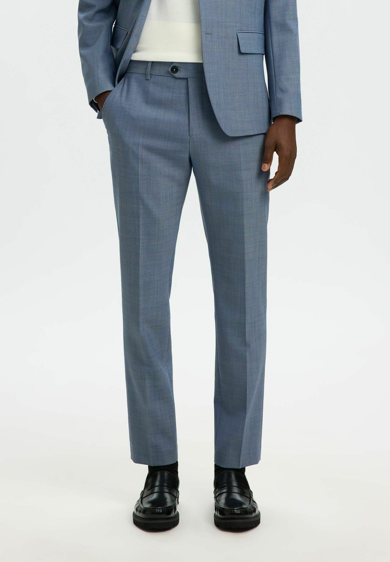 Pantalon bleu clair sur mesure en tissu texturé, avec un devant plat, des poches latérales, porté avec des chaussures habillées noires.