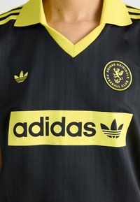 Melns sporta krekls ar dzeltenu apkakli un akcentiem. Iekļauj vertikālas svītras, "adidas" logo dzeltenā krāsā un noapaļotu emblemu uz krūšdaļas.