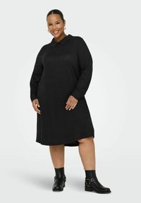 Černé šaty s dlouhým rukávem a límečkem, volný střih, ke kolenům, vyrobené z měkké tkaniny, v kombinaci s černými kotníkovými botami s dekorativními prvky.